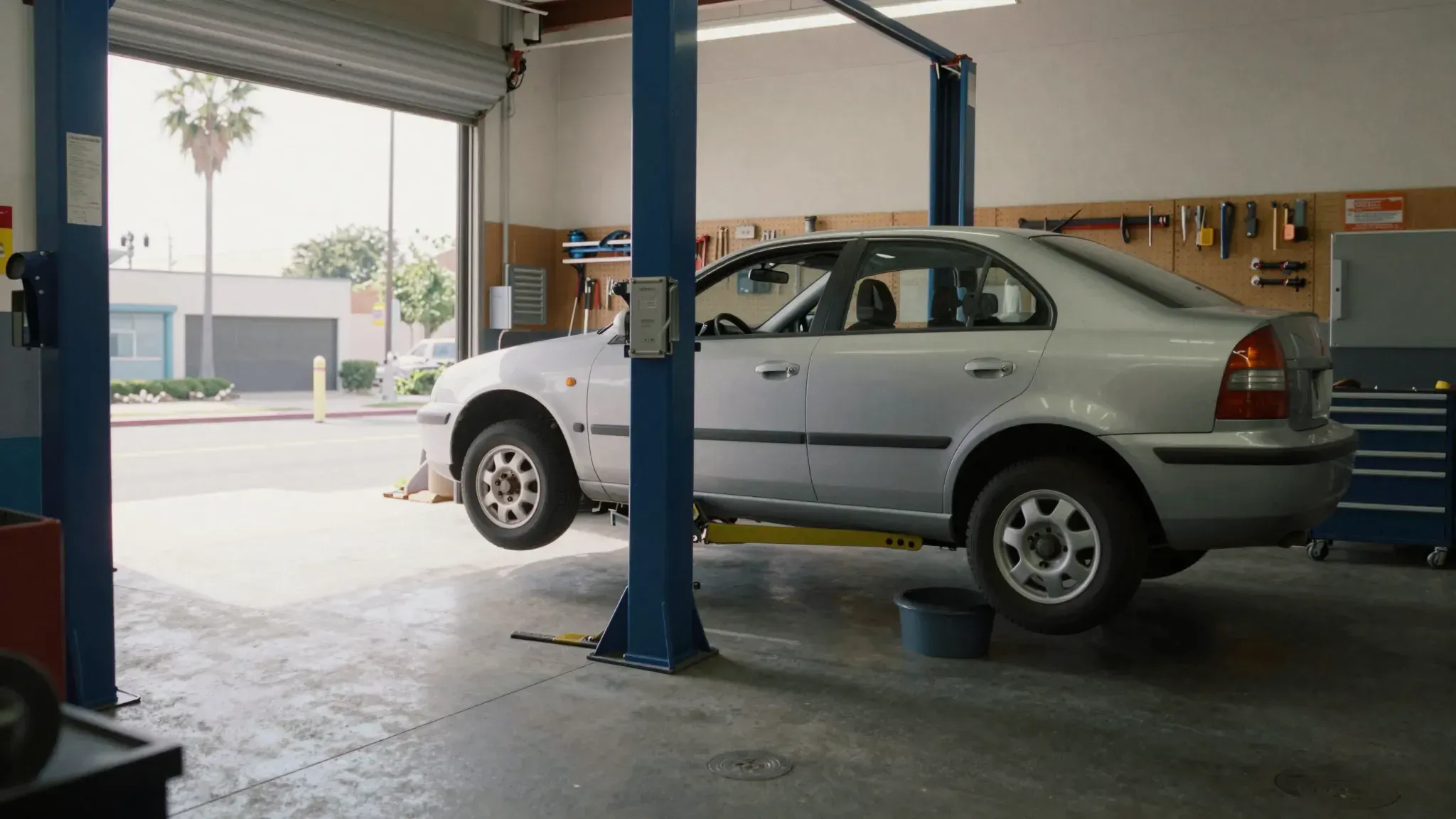 A silver compact car raised on a lift in a clean, orderly repair bay with a palm tree outside.